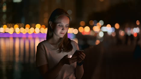 Una-Hermosa-Joven-En-Verano-En-Una-Gran-Ciudad-Nocturna-Mantiene-Un-Teléfono-Inteligente-En-Su-Interior-Y-Escribe-Un-Mensaje-Lee-Correos-Electrónicos-Y-Se-Comunica-En-Salas-De-Chat-Y-Redes-Sociales-Mantiene-Un-Blog-Mira-La-Pantalla-Del-Teléfono